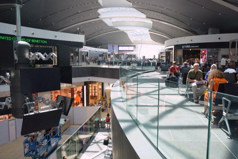 Interno dell'aeroporto con negozi, posti a sedere e passeggeri sotto un tetto curvo con luce naturale.