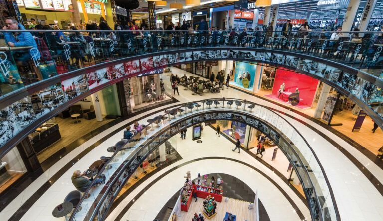 Interno di un centro commerciale a più livelli, con negozi su ogni piano, gente che curiosa, illuminazione intensa.