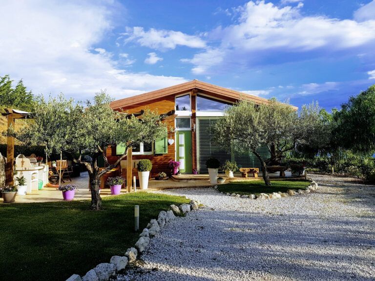 Casa in legno con persiane verdi, ulivi e vialetto di ghiaia sotto un cielo nuvoloso.