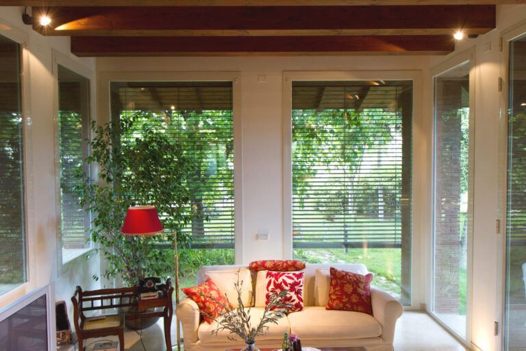 Veranda con divano bianco, cuscini rossi e grandi finestre con vista sugli alberi.