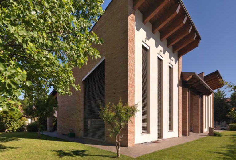 Moderno edificio in mattoni e bianco, con alte finestre e tetto spiovente, circondato da alberi ed erba.