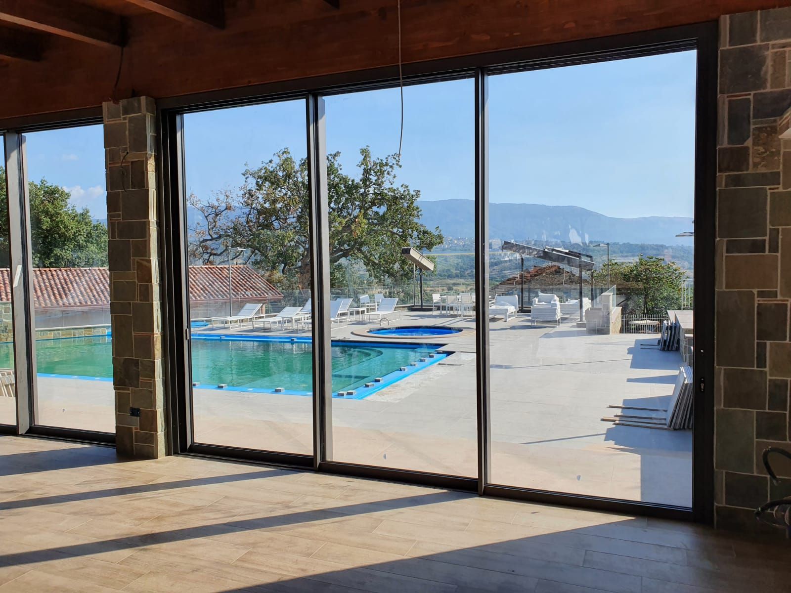 Ampie finestre con vista sulla piscina e sul patio. Interni dell'edificio con pavimento piastrellato e pilastri in pietra.