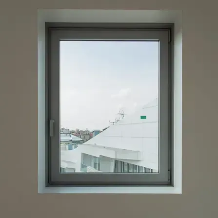 Finestra che incornicia la vista del tetto di un edificio bianco e di un paesaggio urbano sotto un cielo azzurro pallido.