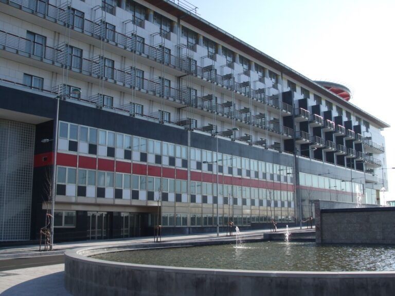Edificio moderno con fasce nere, rosse e bianche, balconi e una fontana circolare in primo piano.