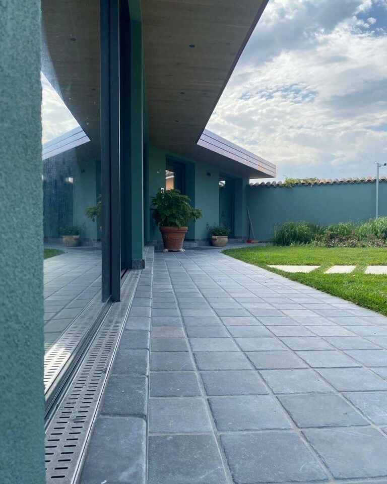 Vista esterna di un edificio color verde acqua con porte in vetro e un vialetto pavimentato che conduce a un giardino.
