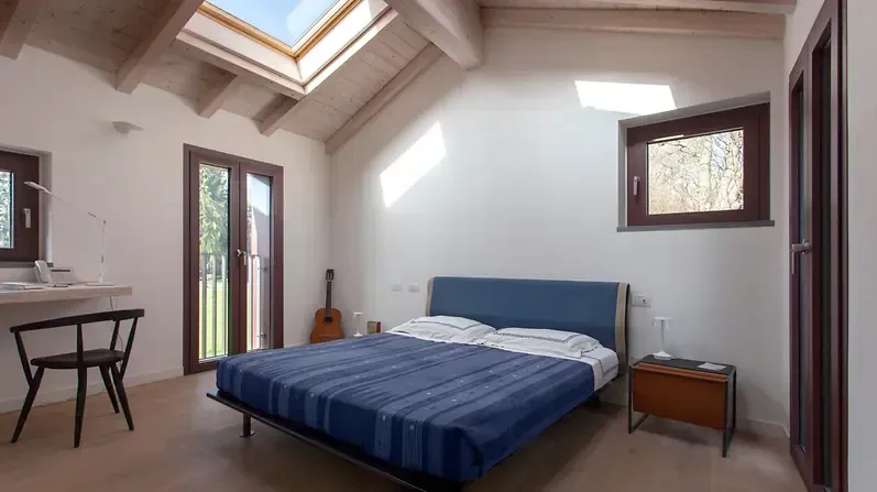 Camera da letto con letto blu, scrivania con sedia, finestre e lucernario; pareti bianche, travi in legno.