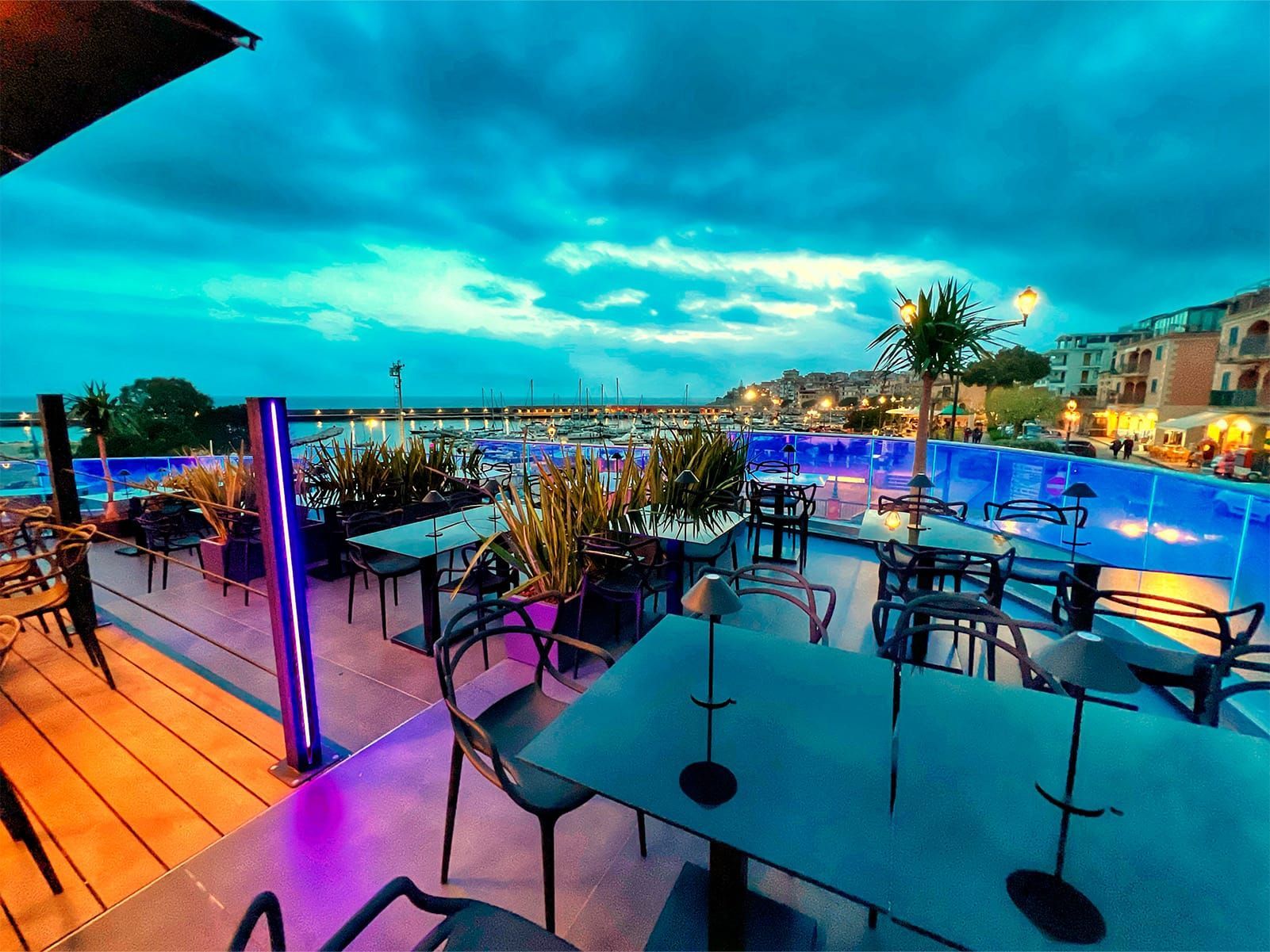 Ristorante all'aperto con tavoli, sedie, piante e giochi d'acqua, con vista sul porto al tramonto e illuminazione blu.