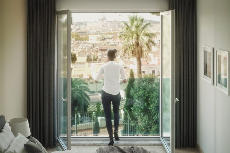 Una donna in camicia bianca e jeans guarda da un balcone una città con le palme.