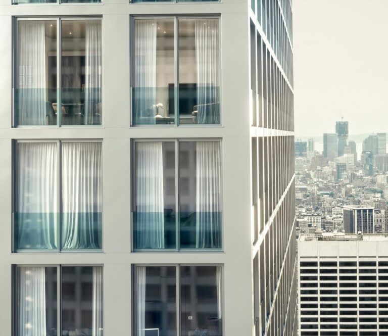 Moderno edificio alto con ampie finestre che si affacciano sullo skyline della città. Le finestre sono dotate di tende bianche.