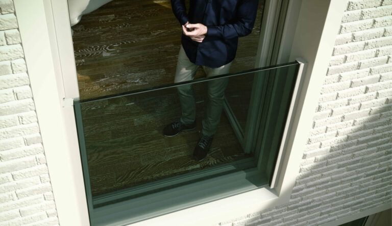 Persona in piedi vicino a una finestra aperta. Gambe e mani visibili; è visibile anche l'esterno dell'edificio in mattoni.