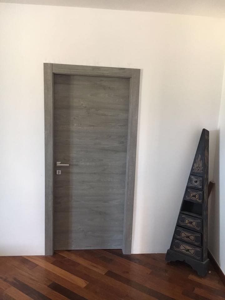 Porta in legno grigio con telaio coordinato, inserita in pareti bianche, pavimento in legno e un alto elemento decorativo.