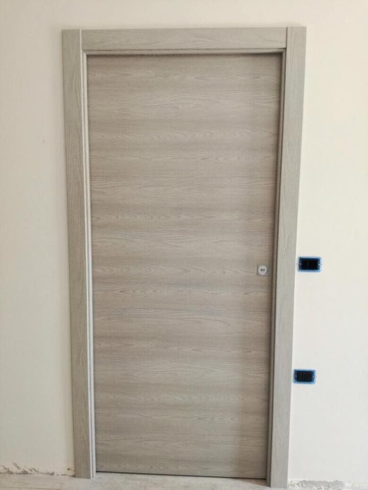 Porta in legno chiaro con cornice coordinata su parete bianca, design semplice. Due copripresa scuri a destra.