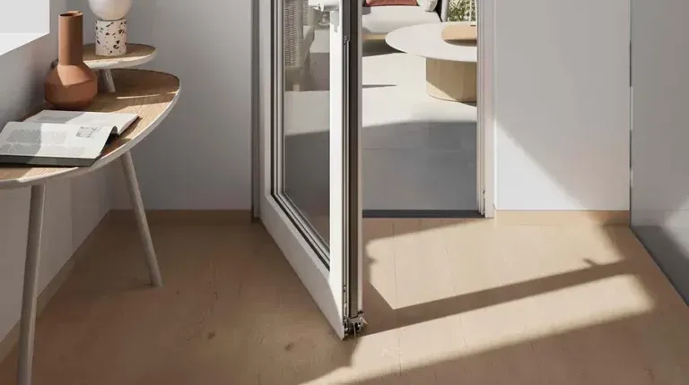 Vista interna con porta aperta, pavimenti in legno chiaro e tavolo con oggetti. Luce solare intensa.
