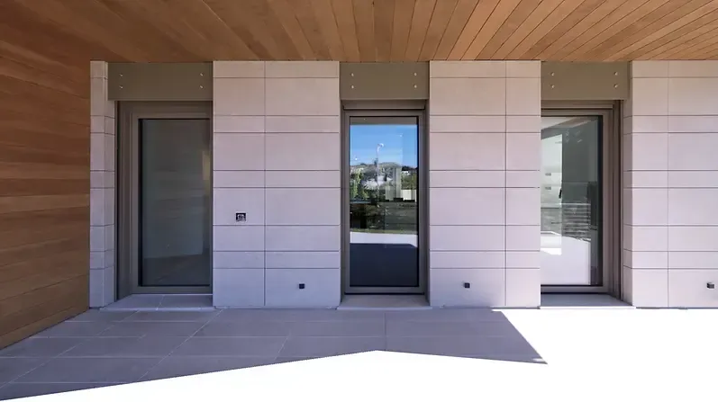 Vista esterna di un edificio con tre porte in vetro, pareti piastrellate bianche e soffitto in legno.