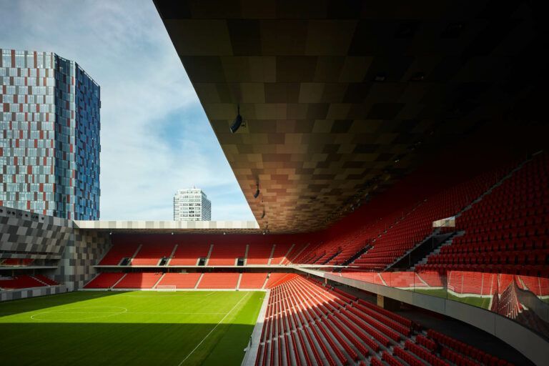 Vista dai posti a sedere dello stadio verso il campo verde e gli alti edifici; posti a sedere rossi, tetto parziale, cielo azzurro.