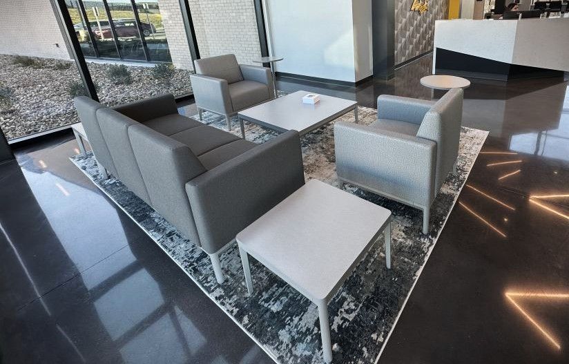 Modern office lounge with gray chairs, small tables, and a patterned rug near large windows