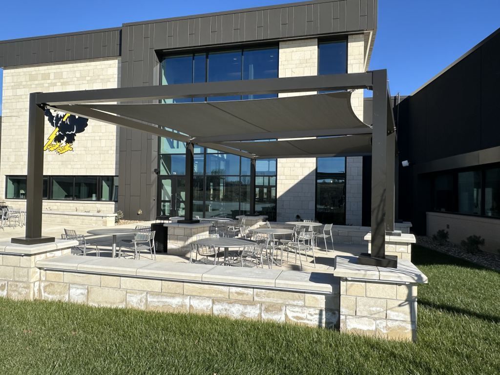 Modern building entrance with a stone patio, pergola frame, and outdoor seating under a clear blue sky