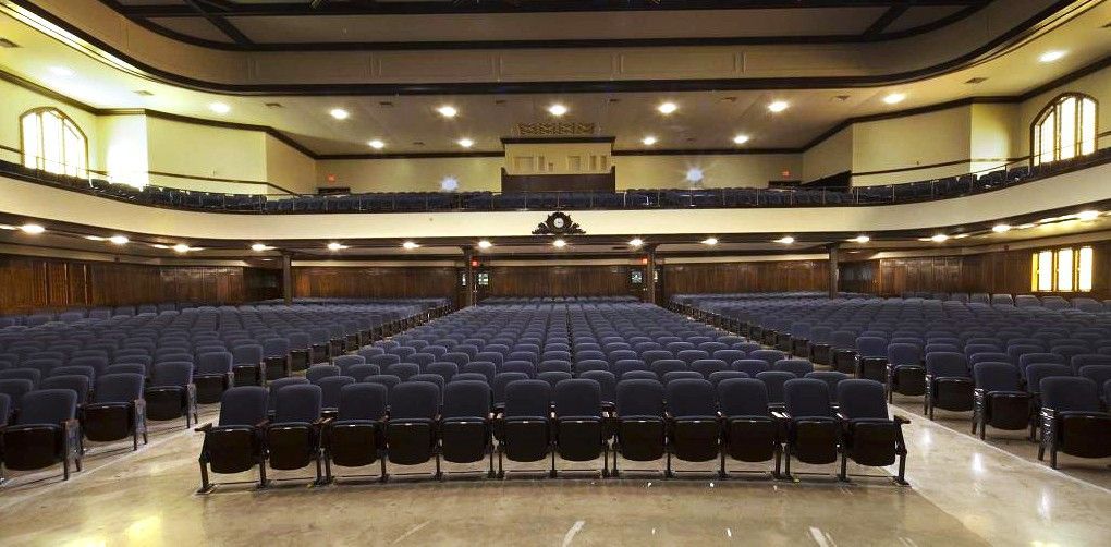 Empty auditorium with rows of blue seats and a stage, viewed from the back toward the screen.