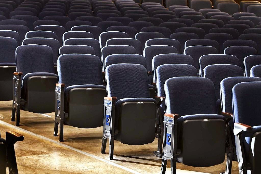 Rows of empty dark auditorium seats with light wood armrests in a theater or lecture hall.