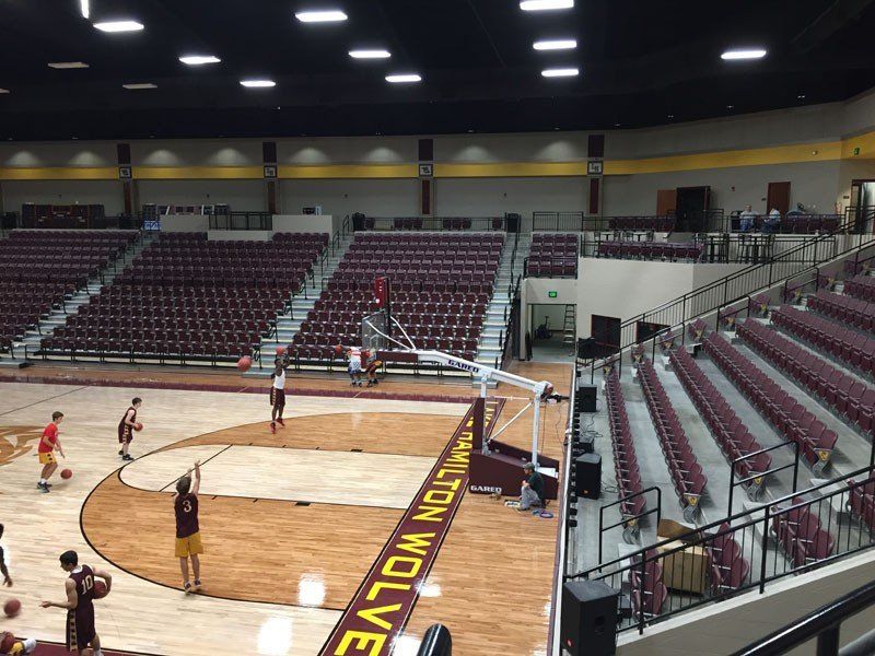Basketball game on a maroon-and-wood court inside an empty arena with stands and overhead lights.