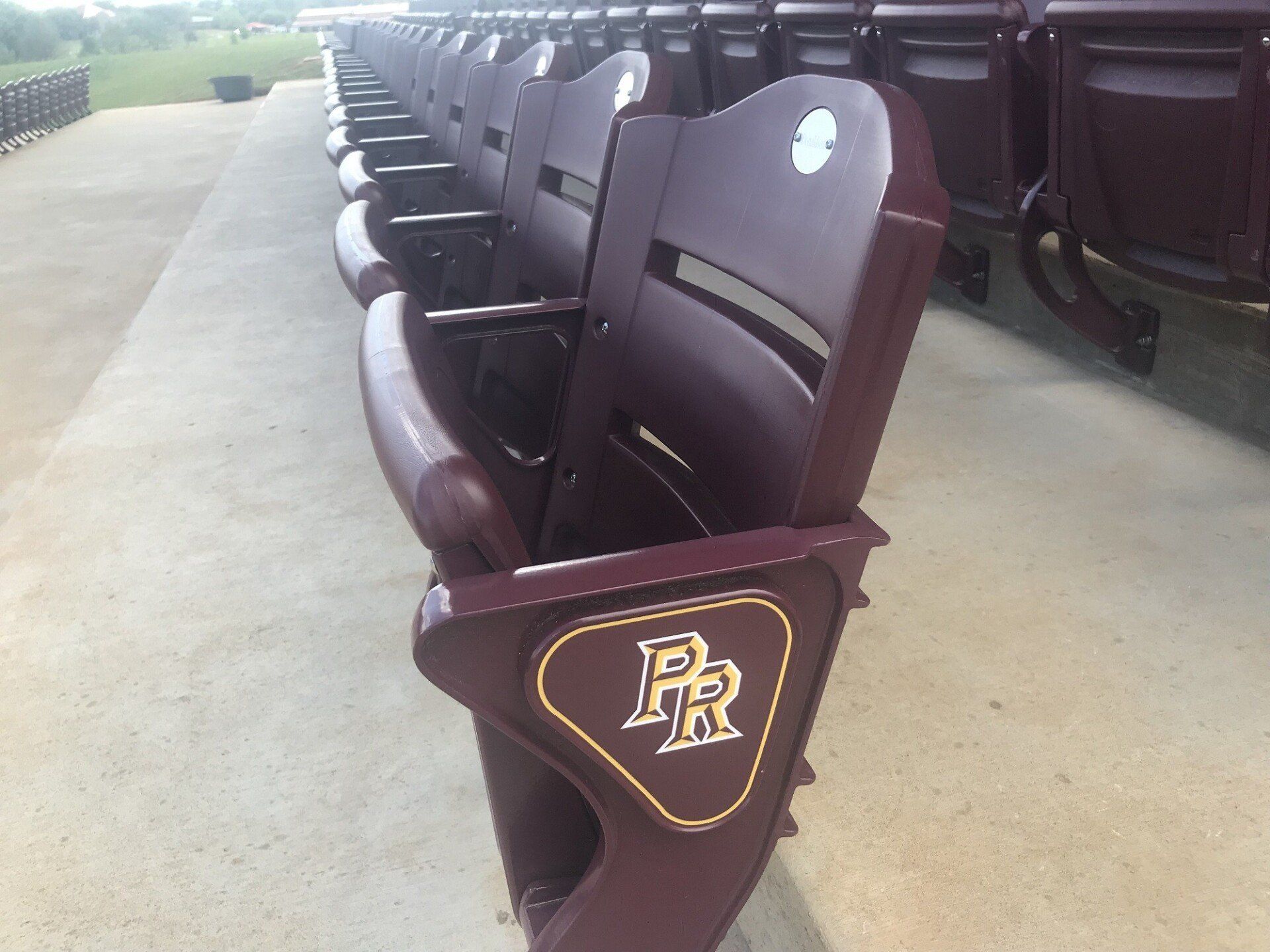 Rows of maroon stadium seats with a gold PR logo on the aisle seat divider, viewed from the side.