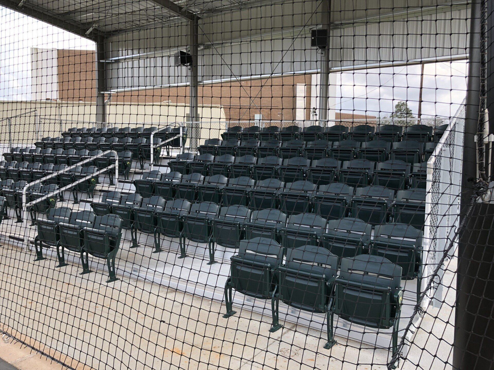 Empty outdoor bleachers under a covered roof, viewed through a chain-link fence.
