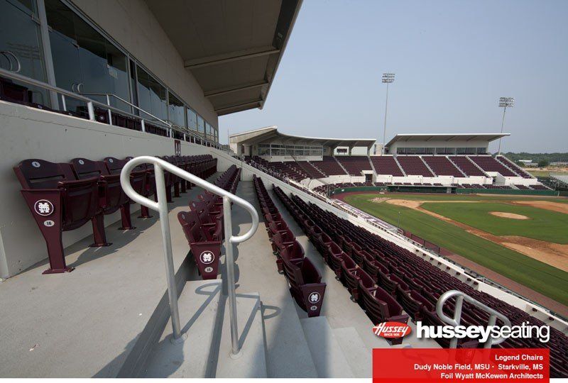 Empty stadium seating overlooking a baseball field, with maroon seats and a white railing.