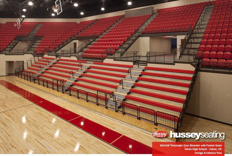 Indoor arena bleachers with red and white seating and polished wood floor, shown from the court side
