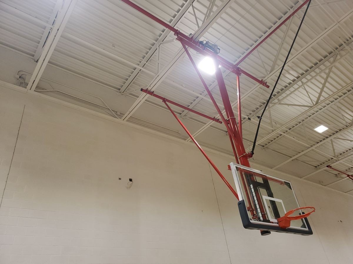 Gym basketball hoop with red support and white backboard mounted in a high ceiling gymnasium