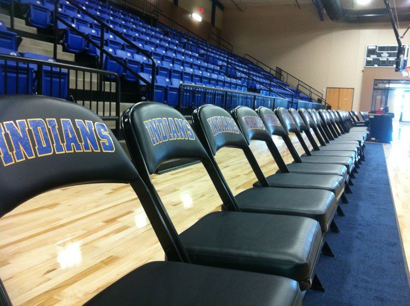 Empty gym bleachers beside a basketball court, with blue seats and black folding chairs on the sideline