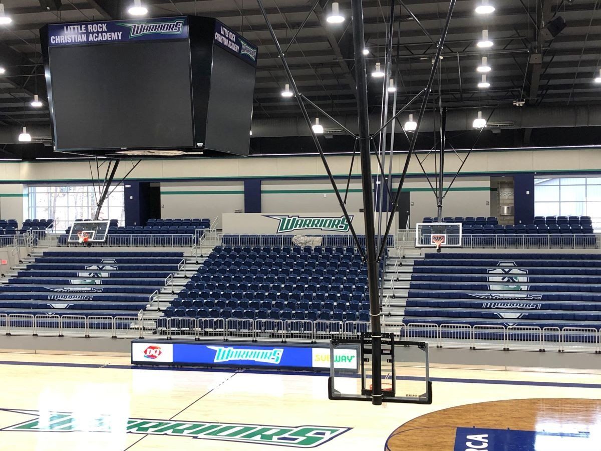 Empty indoor basketball arena with blue bleachers, a hanging scoreboard, and hoops at center court