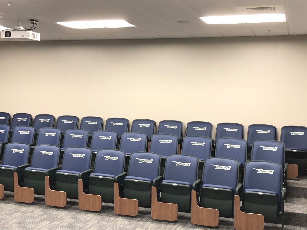 Empty lecture hall with rows of blue seats and a projector on the ceiling.