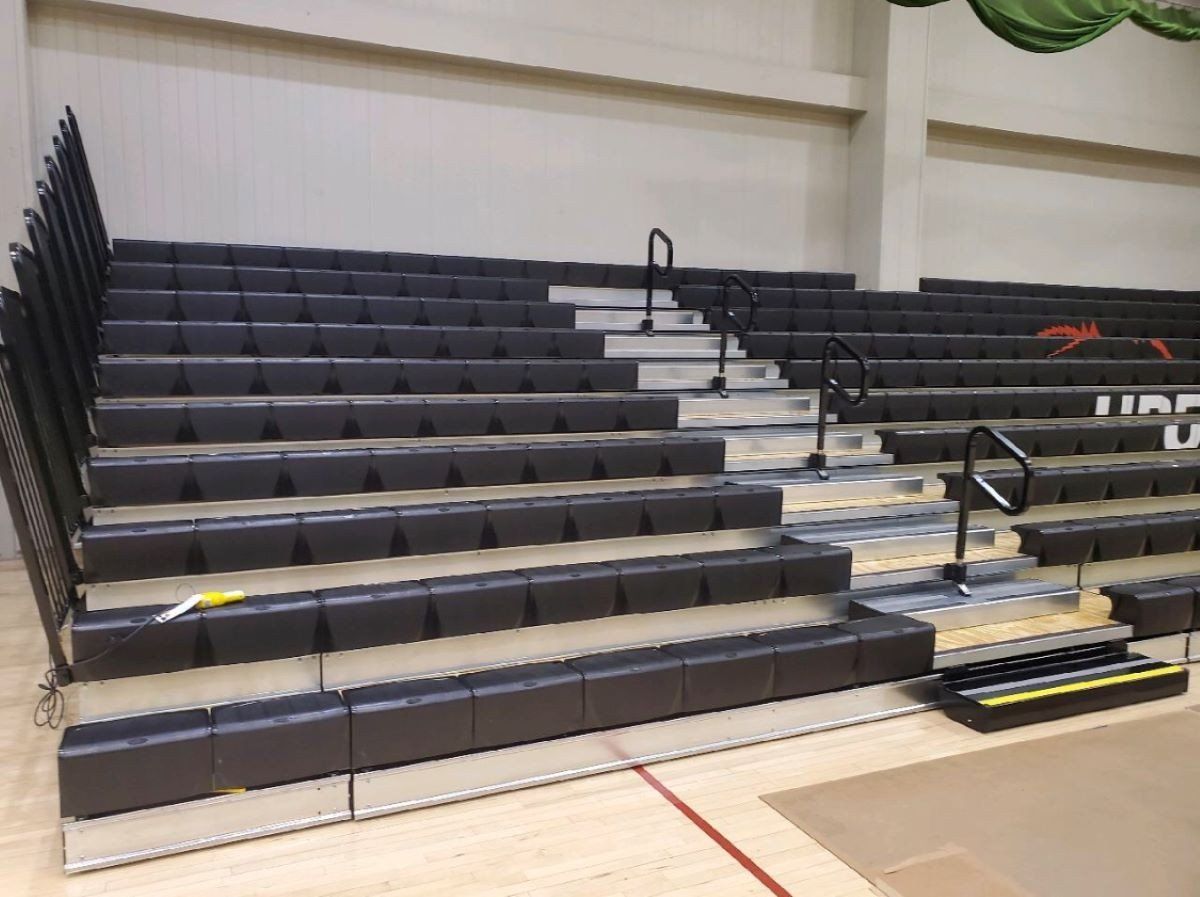 Indoor bleachers with black folding seats in a gymnasium, shown empty and folded up.