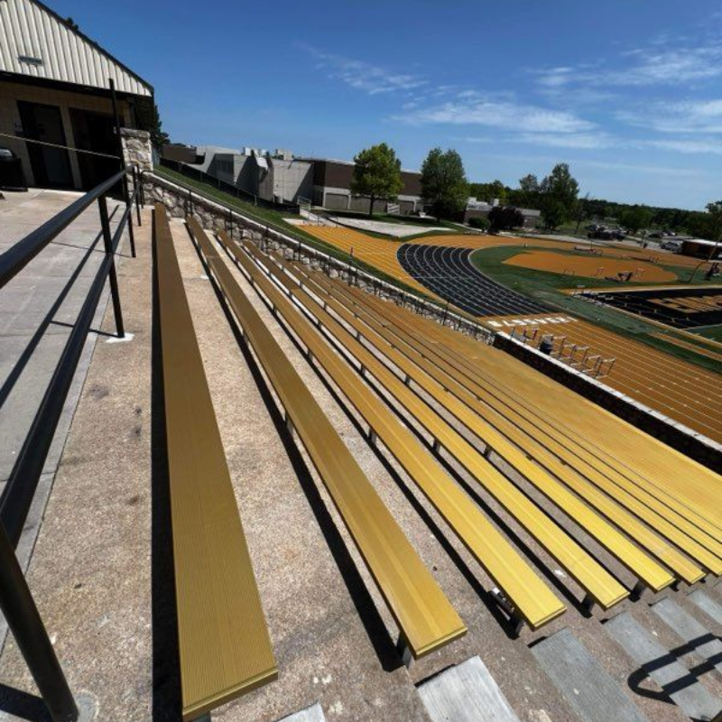 Rows of yellow bleachers beside a sports field under bright sunlight, with a stadium building at left.