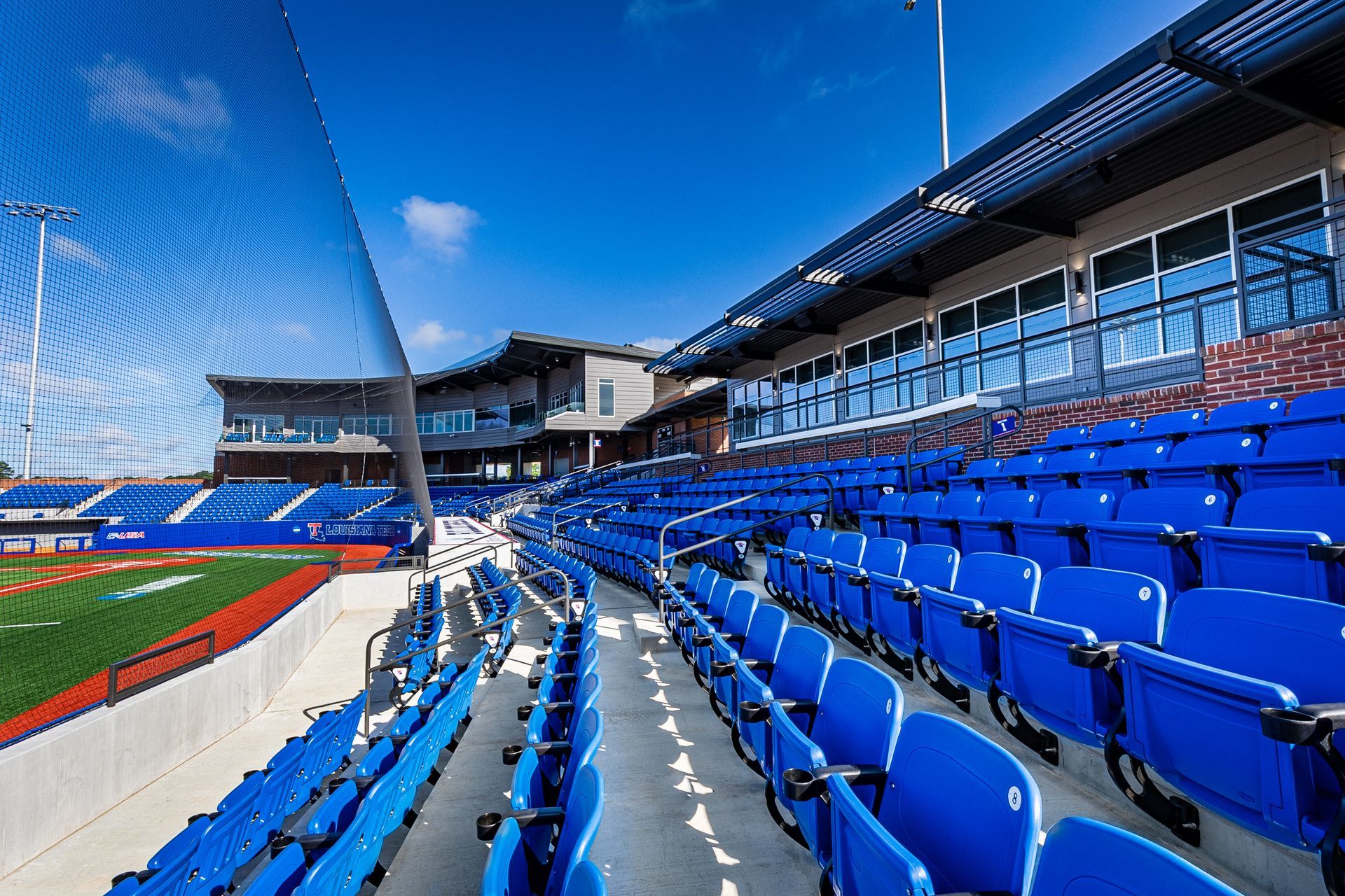 Empty stadium seating with bright blue chairs beside a running track and field under a clear sky