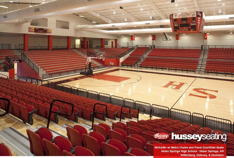 Empty indoor basketball arena with red seats and a polished court, featuring Hussey Seating branding.