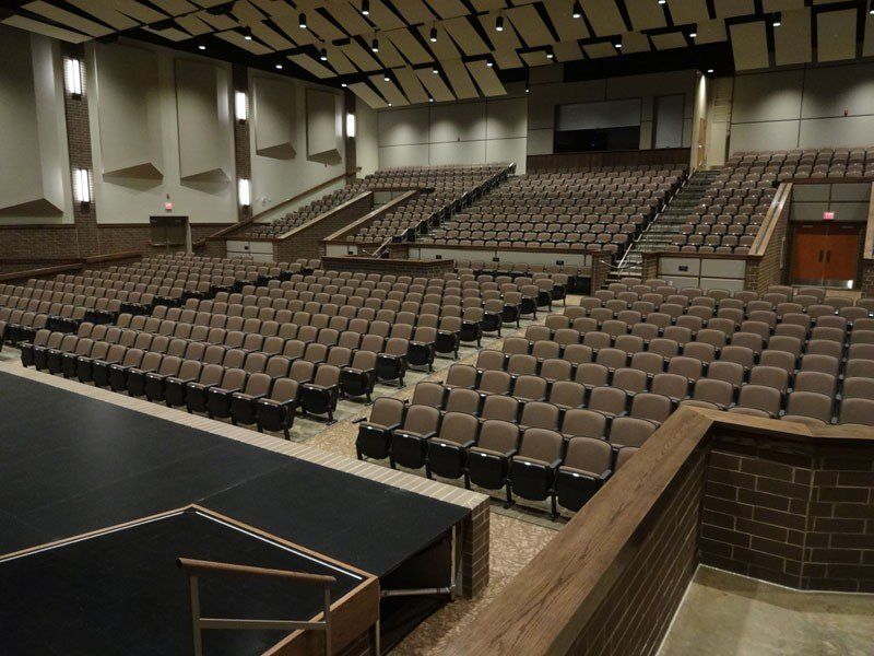 Empty auditorium with rows of tan seats, stage at left, and brick walls under a high ceiling
