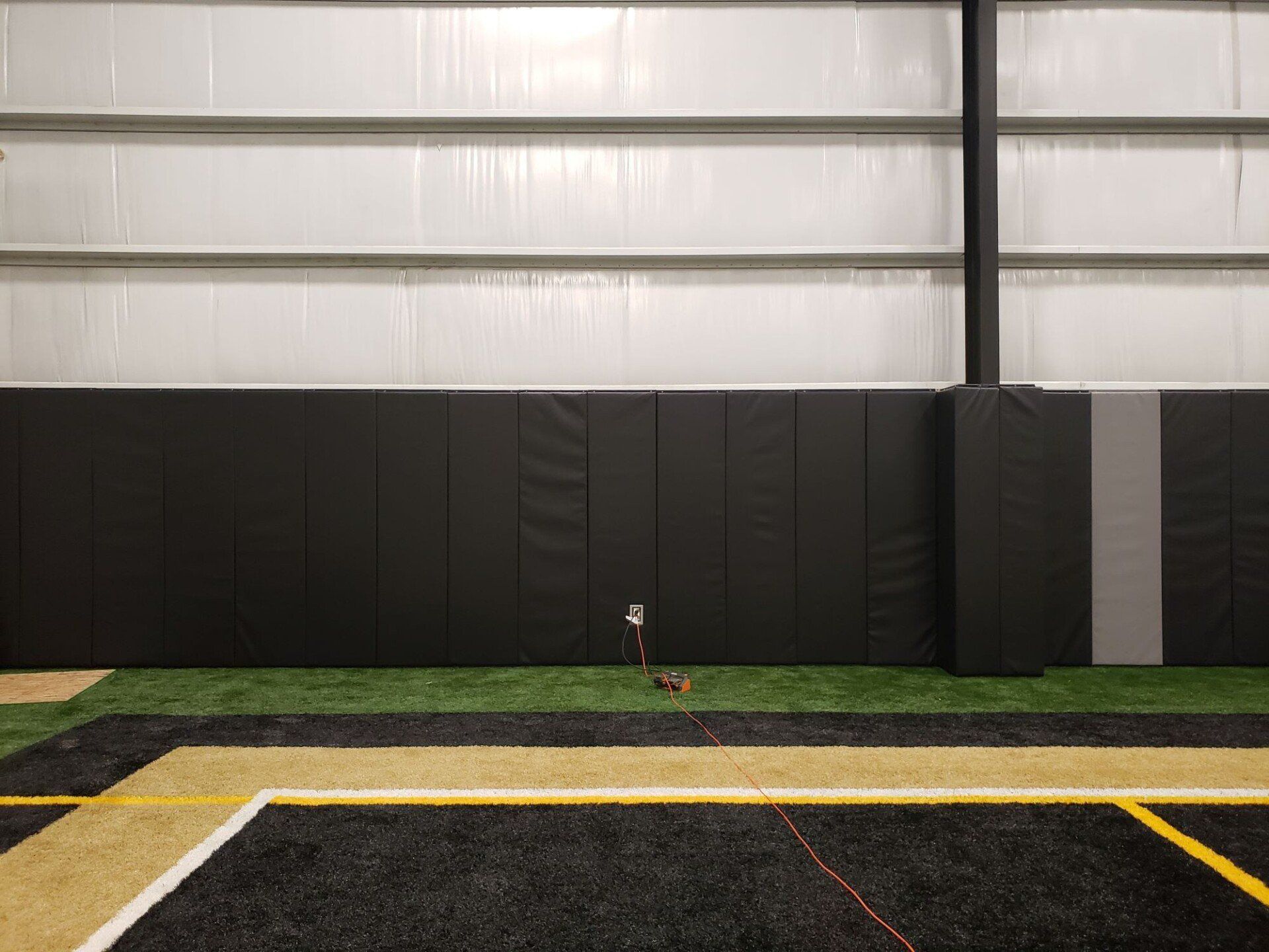 Indoor sports field with black padded wall, green turf, and yellow boundary lines.