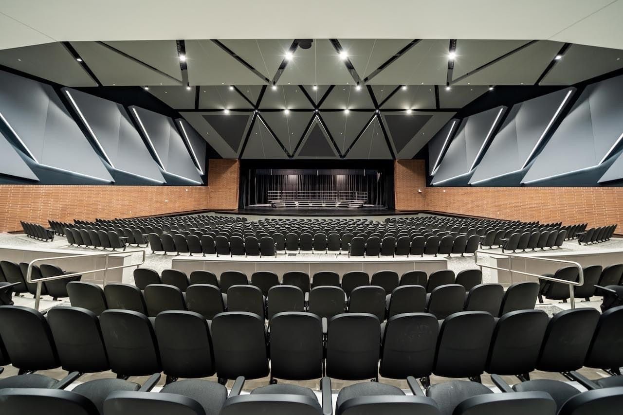 Empty auditorium with rows of black seats and a stage under geometric ceiling lights