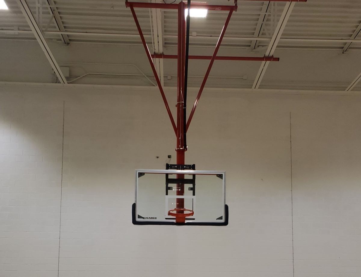 Basketball hoop and backboard hanging from the ceiling in an indoor gym.