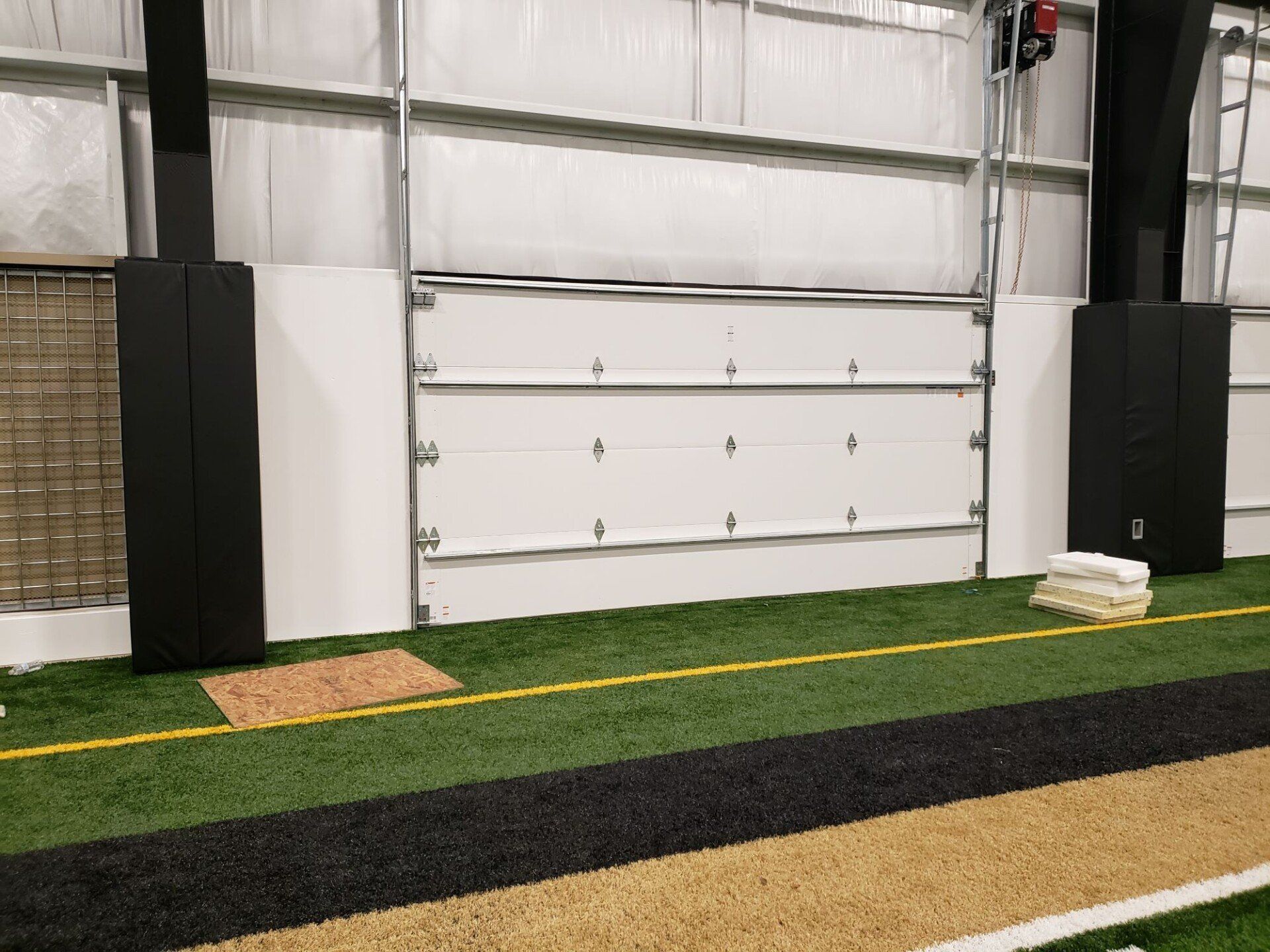 Indoor turf training area with a garage door, black wall pads, and yellow boundary lines