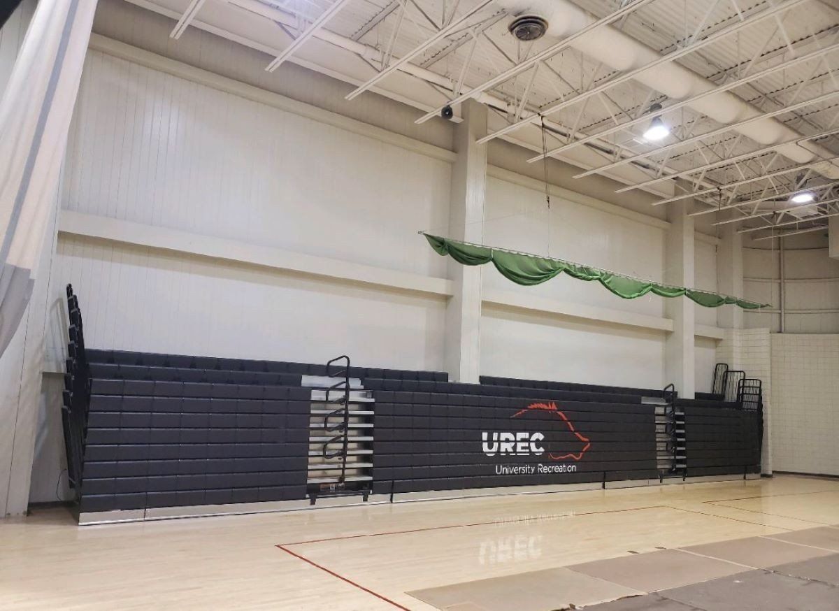 Indoor gymnasium with stacked dark blue bleachers, a ladder, and a hanging green net near the ceiling.