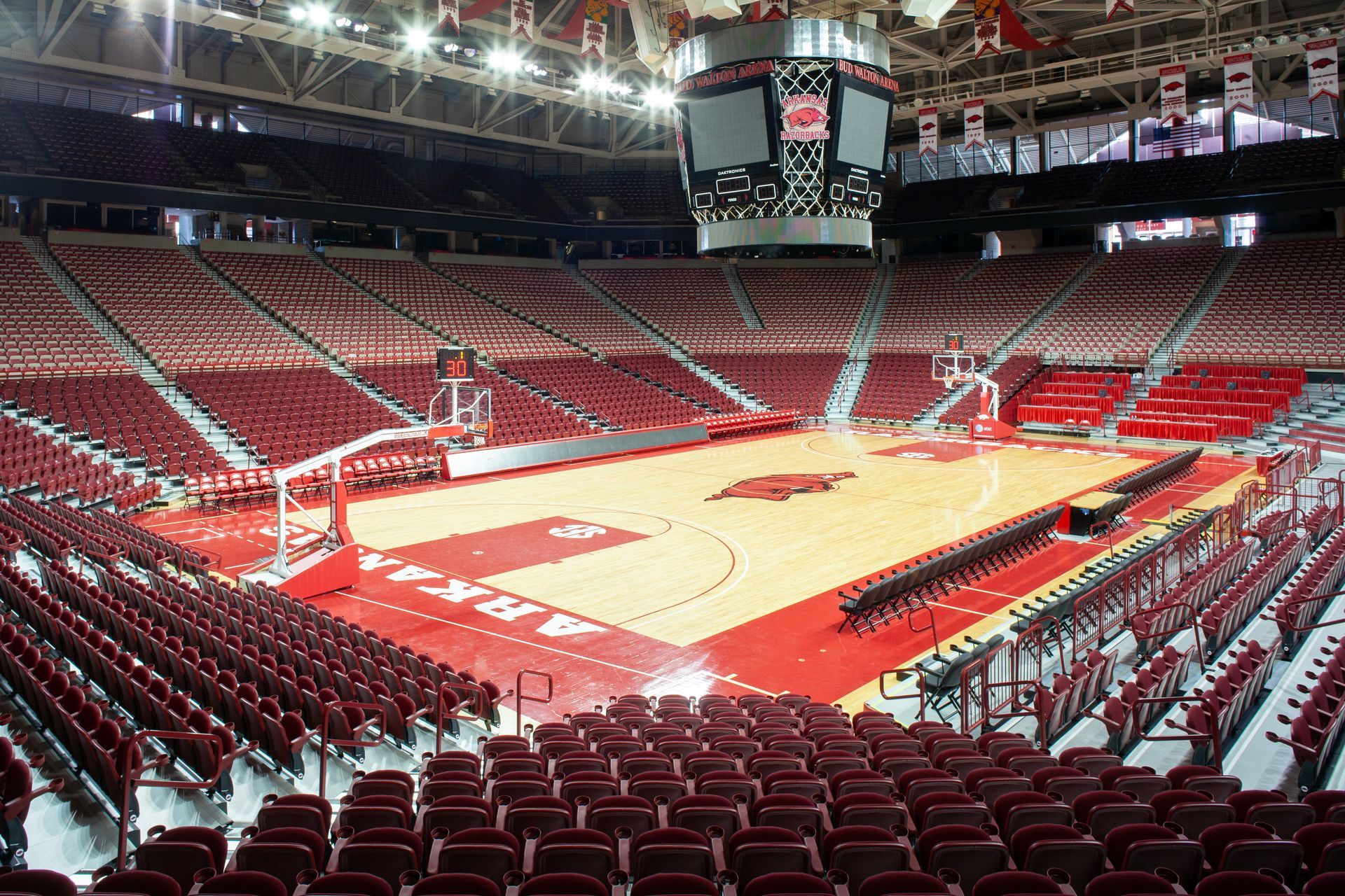 An empty basketball arena with red seats and a hardwood court featuring a large Arkansas Razorbacks logo in the center.