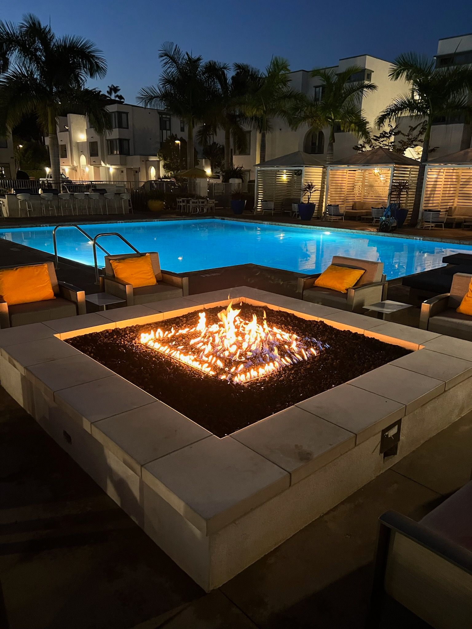 A fire pit is lit up in front of a swimming pool at night.