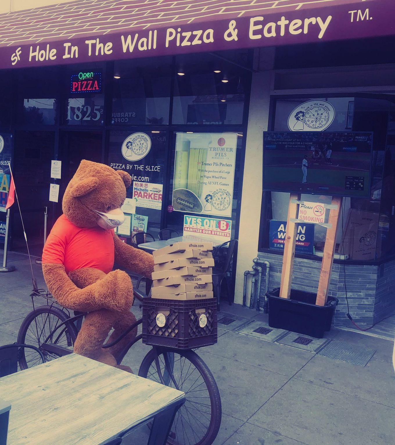 A teddy bear is riding a bike in front of a hole in the wall pizza & eatery