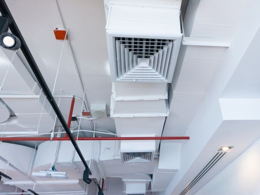 Ceiling view of white HVAC vents, exposed pipes, and red sprinkler line in an industrial interior