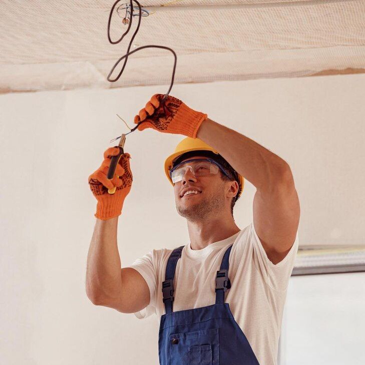 Electrician in blue overalls and orange gloves using tools to work on ceiling wiring