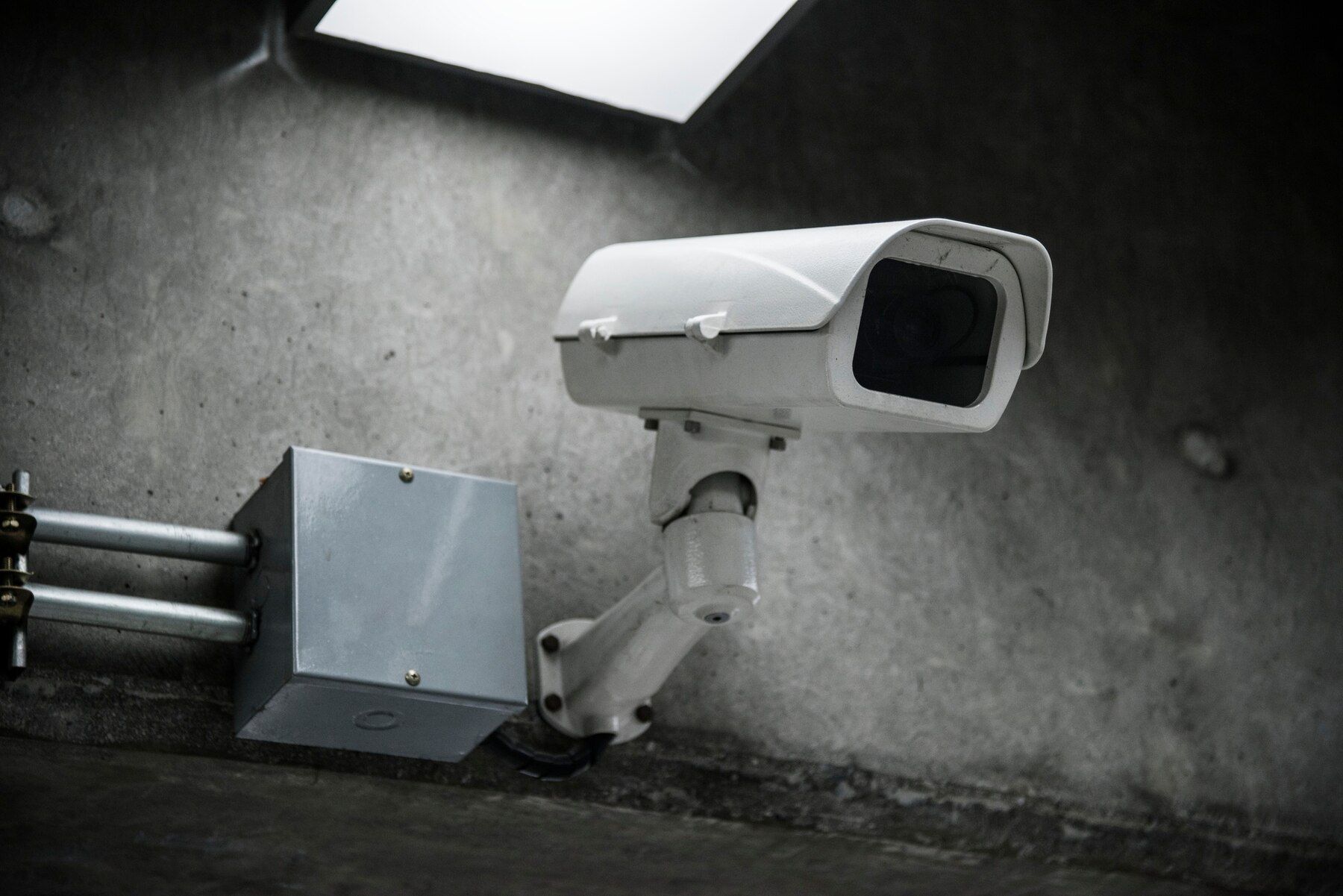 White security camera mounted on a concrete wall beside pipes in a dim industrial setting