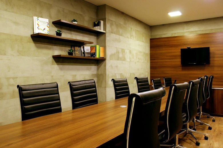 Modern conference room with a long wooden table, black chairs, wall shelves, and a wall-mounted TV.