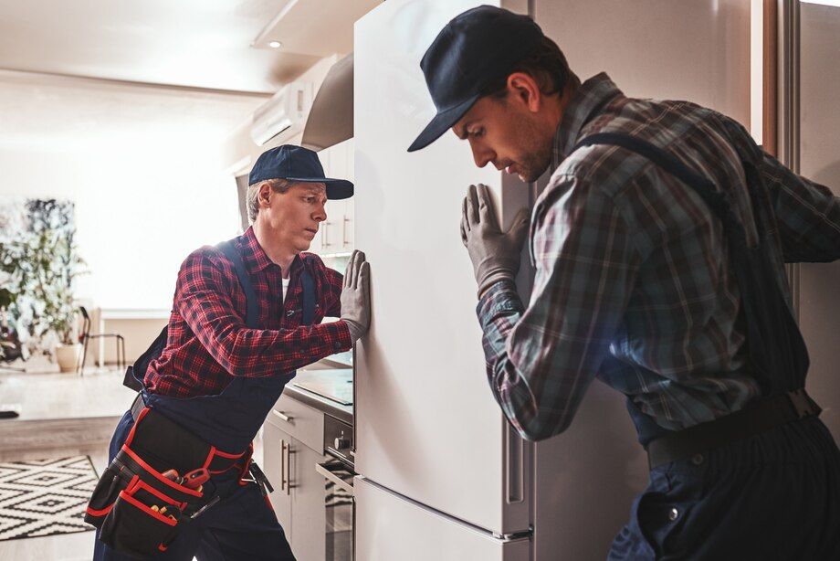 Two workers in caps and plaid shirts move a large appliance through a bright kitchen doorway.
